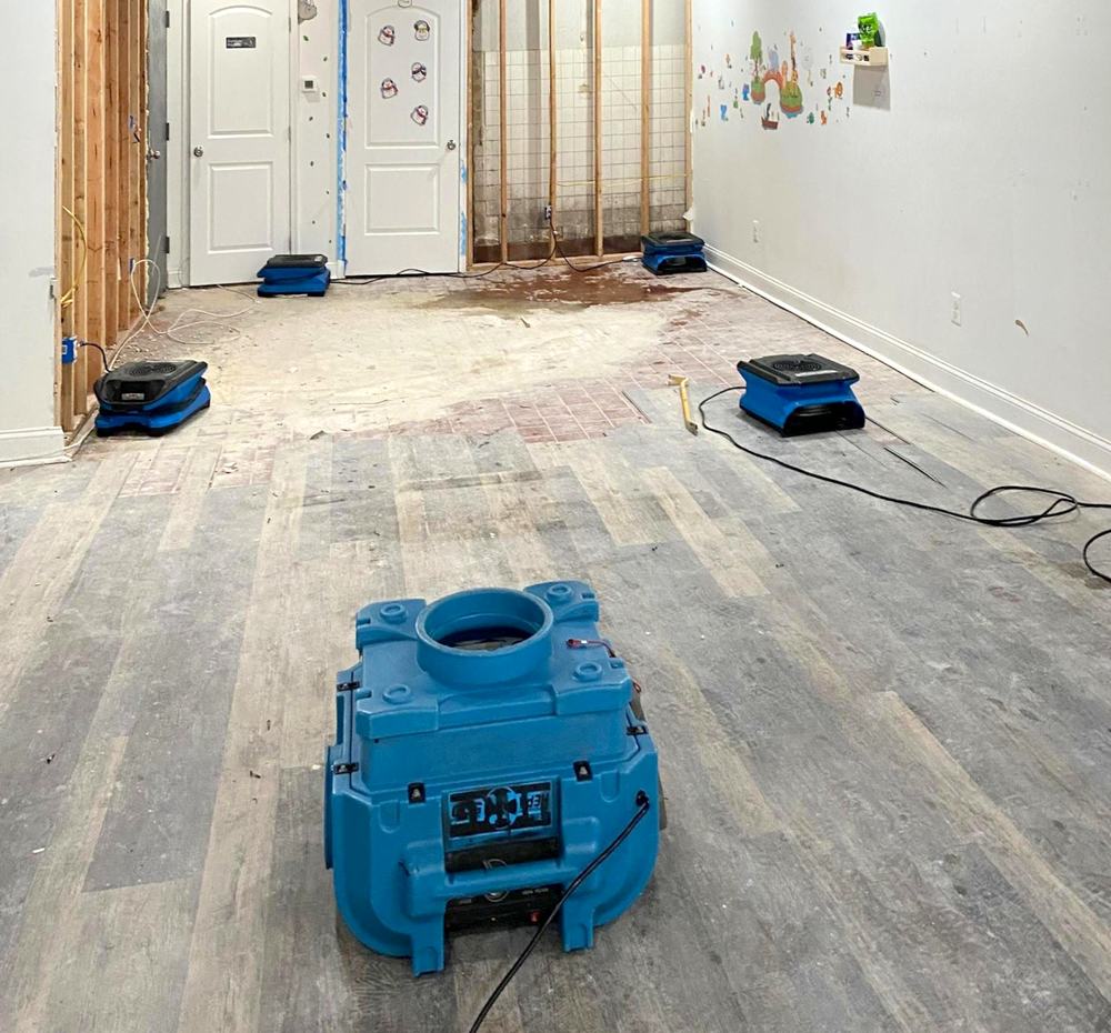 Room with part of a wall opened to the studs after water damage, with a blue air scrubber in the foreground and air movers running along the walls.